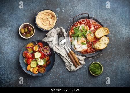 Traditionelles nahöstliches Frühstück oder Brunch mit Eiern, Shakshouka in der Pfanne mit Toast, frischem Gemüsesalat, Hummus und Oliven auf rustikalem Beton Stockfoto