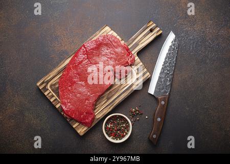 Rohes rohes, rohes, rundes Rindersteak auf Holzschneidebrett mit großem Küchenmesser und Pfeffer auf dunkelbraunem rustikalem Steinhintergrund, Blick von oben, kochte mich Stockfoto