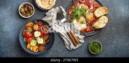 Traditionelles nahöstliches Frühstück oder Brunch mit Eiern, Shakshouka in der Pfanne mit Toast, frischem Gemüsesalat, Hummus und Oliven auf rustikalem Beton Stockfoto