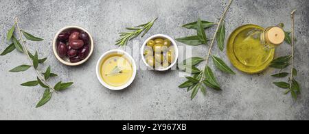 Zusammensetzung mit grünen und braunen Oliven, extra natives Olivenöl in Glasflasche, Olivenbaumzweige auf grauem Betonstein rustikalen Hintergrund über dem Kopf Stockfoto