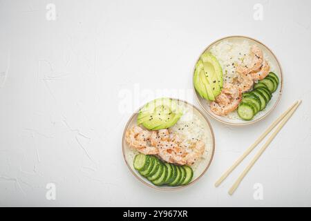 Zwei weiße Keramikschalen mit Reis, Garnelen, Avocado, Gemüse und Sesamsamen und Holzstöcken auf weißem Betonsteinhintergrund von oben. Gesundheit Stockfoto