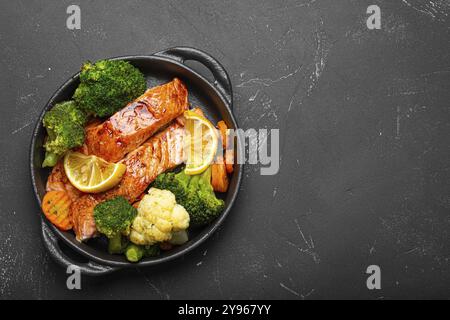 Gesunde gebackene Fisch Lachs Steaks, Brokkoli, Blumenkohl, Karotten in gusseiserner Auflaufschüssel schwarzer dunkler Stein Hintergrund. Ein köstliches, kohlenhydratarmes Essen Stockfoto