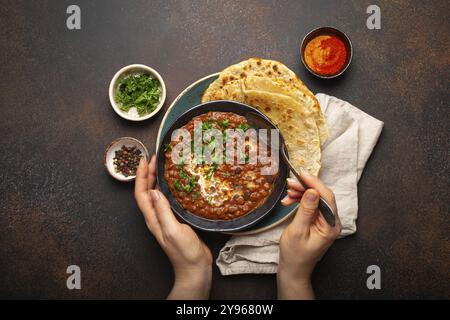 Weibliche Hände halten eine Schüssel und essen traditionelles indisches Punjabi-Gericht Dal makhani mit Linsen und Bohnen, serviert mit Naan-Fladenbrot, frischem Koriander Stockfoto