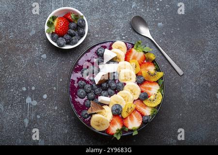 Gesunder Sommer Acai Smoothie Bowl mit Chiasamen, frischer Banane, Erdbeere, Heidelbeere, kokos, kiwi-Blick von oben auf rustikalem Betonhintergrund mit Löffel Stockfoto