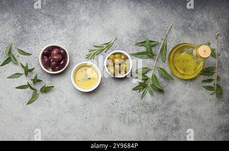 Zusammensetzung mit grünen und braunen Oliven, extra natives Olivenöl in Glasflasche, Olivenbaumzweige auf grauem Betonstein rustikalen Hintergrund über dem Kopf Stockfoto