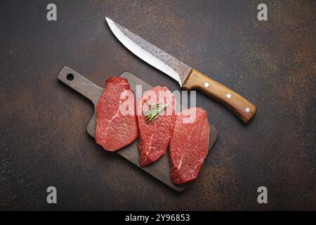 Drei rohe rohe rohe Rindersteaks auf Holzschneidebrett mit Rosmarin auf Holzschneidebrett mit Messer auf dunklem rustikalem Hintergrund Stockfoto