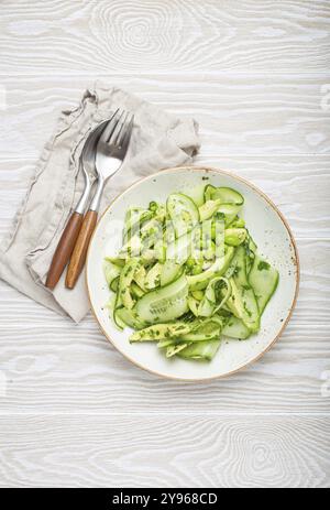 Gesunde vegane grüne Avocadosalatschale mit geschnittenen Gurken, Edamame Bohnen, Olivenöl und Kräutern auf Keramikplatte, Draufsicht auf weißem rustikalen Holztabl Stockfoto