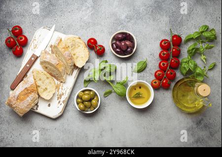 Lebensmittelzusammensetzung mit Ciabatta in Scheiben, Oliven, Olivenöl, Spaghetti, frischem Basilikum, Kirschtomaten auf grauem Betonstein rustikaler Hintergrund von oben. I Stockfoto