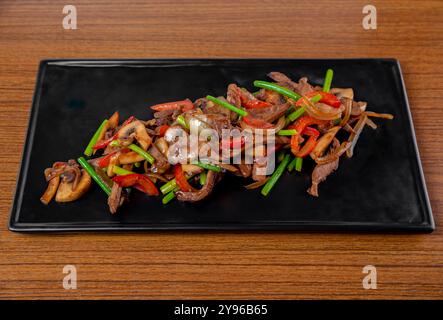 Gebratenes Schweinefleisch und Rindfleisch mit Knoblauchpfeilen, Pilzen und Gemüse auf einem schwarzen Teller auf einem Holztisch. Stockfoto