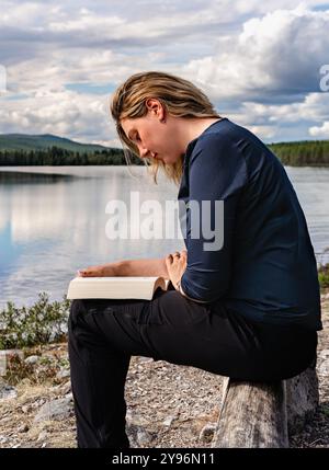 Die schöne Frau sitzt auf einer Holzbank an einem ruhigen See in Schweden, tief vertieft beim Lesen ihres Buches, umgeben von Natur und ruhigem Wasser Stockfoto