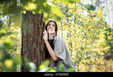 Eine Frau lehnt sich an einen Baum, blickt mit einem ruhigen Ausdruck nach oben, umgeben von leuchtend grünen Blättern in einem sonnendurchfluteten Wald. Stockfoto