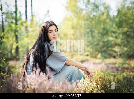 Eine junge Frau sitzt ruhig auf dem Waldboden, ihr langes Haar zieht sich über den Rücken. Sie erscheint tief in Gedanken, umgeben von blühenden Wildblumen Stockfoto