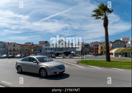 Costa Nova, Portugal - 11. September 2024 : Ein silbernes Fahrzeug fährt an charmanten Gebäuden und Palmen unter hellem Himmel vorbei. Stockfoto