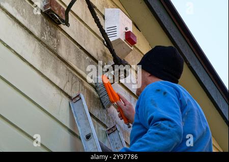 Ein Mann schrubbt ein Haus, bevor er malt. Er ist in der Nähe des elektrischen Eingangs zum Haus und einer Alarmbox Stockfoto