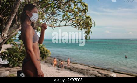 Eine Frau mit Gesichtsmaske steht selbstbewusst am Sandstrand, die Wellen streicheln sanft das Ufer hinter sich. Trotz der Pandemie findet sie Trost und Frieden in der Schönheit des Ozeans. Stockfoto