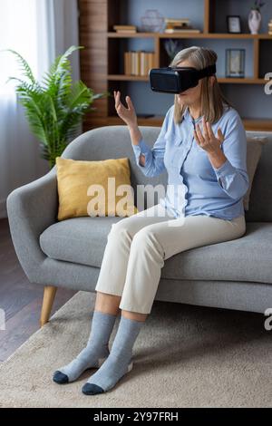 Frau mittleren Alters mit VR-Headset auf der Couch im modernen Wohnzimmer. Erleben Sie virtuelle Realität, entdecken Sie faszinierende Technologien mit Neugier und Spannung in ungezwungener Umgebung. Lifestyle-Konzept. Stockfoto