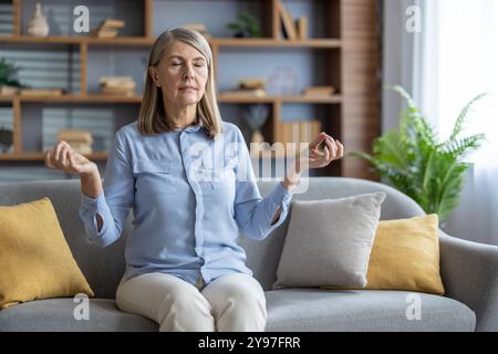 Seniorin meditiert auf Sofa mit ruhigem Ausdruck. Achtsamkeit und Entspannungstechniken zur Stresslinderung in ruhiger häuslicher Umgebung üben. Mit blauem Hemd, bequem sitzend. Stockfoto