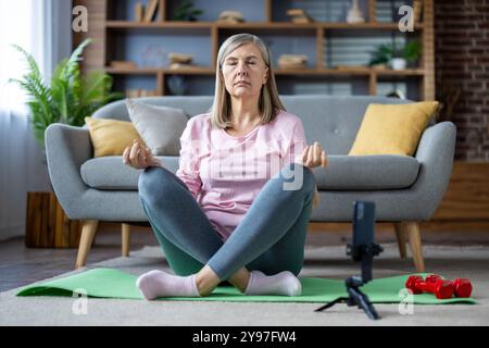 Reife Frau in Lotusposition, die Yoga auf grüner Matte im Wohnzimmer übt, während die Smartphone-Kamera die Sitzung aufzeichnet. Entspannung, Achtsamkeit und ganzheitlicher Wellness-Ansatz zu Hause. Stockfoto