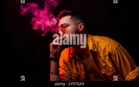 Ein Mann raucht eine zündete Zigarre, der Rauch wirbelt herum und fängt das Feuer ein. Mann in Brille und rauchender Zigarre vor schwarzem Hintergrund. Mann mit Zigarre Stockfoto