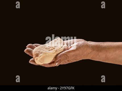 Alte Frauenhand, die einen Kuchen hält, Teig auf schwarzem Hintergrund. Weibliche Hände, die rohen Teig auf schwarzem Hintergrund halten. Hände, die einen fertigen, sauberen Teig halten Stockfoto