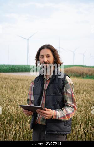 Landwirt mit digitalem Tablet arbeitet auf einem landwirtschaftlichen Feld bei Sonnenuntergang. Windräder im Hintergrund. Stockfoto