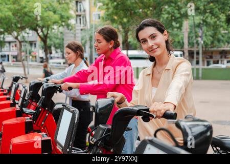 Gruppe junger Frauen, die Leihfahrräder für eine Fahrt in der Stadt nehmen und umweltfreundliche Verkehrsmittel, Outdoor-Aktivitäten und urbane Pendelfahrten mit Schwerpunkt auf Nachhaltigkeit und gesunder Lebensweise präsentieren. Hochwertige Fotos Stockfoto