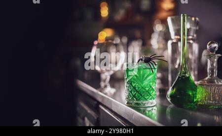 Grüner Halloween-Cocktail in einem Kristallglas mit Spinnendekoration, sitzt auf einem Bartheken neben antiken Glastränkeflaschen, Partyhintergrund Stockfoto