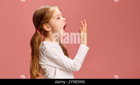 Seitenansicht eines Mädchens, das gähnt und ihre Hand nach vorne streckt, während sie sichtbare Anzeichen von müde oder schläfrig vor pastellrosa Hintergrund zeigt. Stockfoto