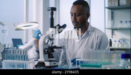Afroamerikanischer Wissenschaftler schaut unter das Mikroskop, untersucht Proben neuer Arzneimittelformel im modernen medizinischen Labor. Im Hintergrund gibt eine Mikrobiologin Daten in den PC ein. Medizinkonzept. Stockfoto