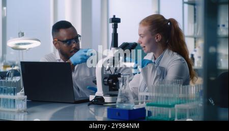 In fortgeschrittenen medizinischen Wissenschaftslabors arbeitet ein vielfältiges Team von zwei Wissenschaftlern an der Entwicklung neuer Medikamente. Afroamerikanischer Mann und kaukasische Frau, die unter dem Mikroskop die Daten auf dem Laptop überprüfen. Stockfoto