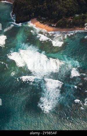 Blick auf brechende Wellen mit türkisfarbenem Wasser an einem Strand mit Palmen, wie von oben gesehen während eines Hubschrauberrundflugs an der Na Pali Coast in Kauai (Hawaii, Stockfoto