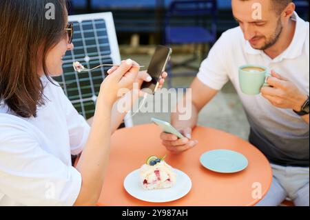 Mann und Frau laden Smartphone vom Sonnenkollektor auf, entspannen im Café. Niedliches Paar, das tragbares Solarladegerät für Geräte verwendet. Charmantes Paar, das auf der Terrasse auf dem Hintergrund einer Solarbatterie sitzt. Stockfoto