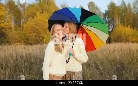 Erwachsene Tochter küsst ihre glückliche, lächelnde, reife Mutter mit buntem Regenschirm, der mit gelben Blättern im Herbstpark spaziert Stockfoto