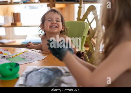 Zwei kleine Kinder sitzen an einem mit Farbe bedeckten Tisch mit einem riesigen Lächeln im Gesicht. Selektiver Fokus Stockfoto