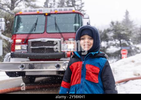 Fröhlicher Junge posiert vor einem roten Feuerwehrauto bei schneebedecktem Wetter und strahlt Urlaubsfreude und Unschuld aus. Warm gekleidet verkörpert er Spaß im Freien und symbolisiert Sicherheit und gemeinnützigen Dienst. Selektiver Fokus Stockfoto