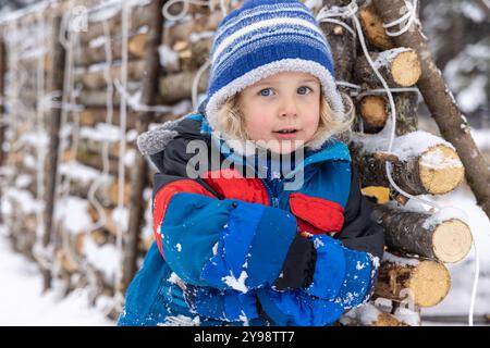 Ein kleiner Junge mit blauem Wintermantel und blau-weißer Haube steht neben einem Stapel Baumstämme im Schnee. Selektiver Fokus Stockfoto