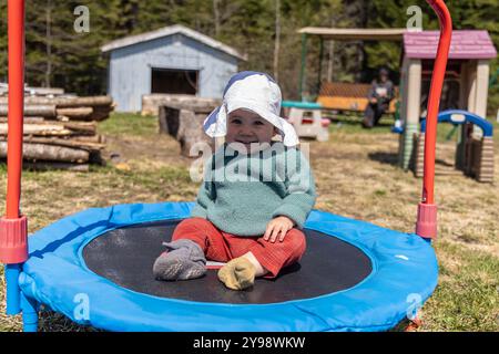 Glückliches Mädchen sitzt auf einem Trampolin im Garten, trägt einen Sonnenhut und lächelt. Selektiver Fokus Stockfoto