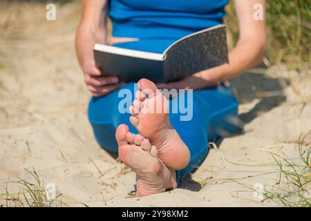 Nahaufnahme einer Frau, die am Strand sitzt und ein Buch liest, Sandfüße Stockfoto