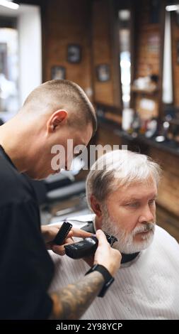 Junger Mann, Friseur, der mit Präzision am Bart des Kunden arbeitet, mit elektrischem Trimmer. Ein älterer Mann besucht den Friseur Stockfoto
