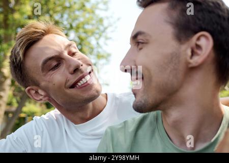 Zwei charmante Männer teilen Lächeln und Lachen, während sie sich in einer lebhaften Parklandschaft umarmen. Stockfoto