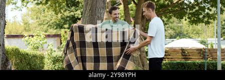 Zwei lächelnde Männer teilen sich einen leichten Moment, während sie eine Decke in einer wunderschönen Umgebung im Freien arrangieren. Stockfoto