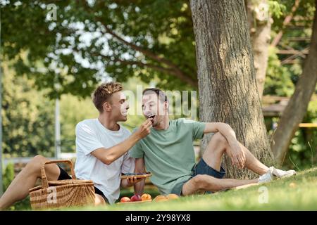 Zwei glückliche Männer genießen ein schönes Picknick und füttern sich an einem warmen Tag unter einem Baum. Stockfoto