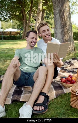 Zwei glückliche Männer teilen sich ein Picknick auf einer Decke und genießen Lachen und Lesen im Freien. Stockfoto