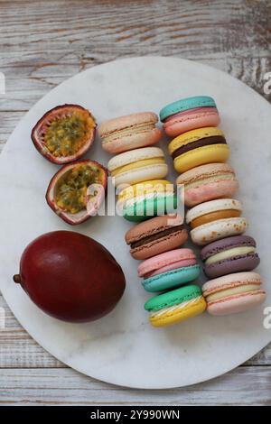 Macarons in verschiedenen Farben und Geschmäckern und Mango- und Passionsfruchthälften auf der Marmorplatte auf dem hölzernen Hintergrund Stockfoto