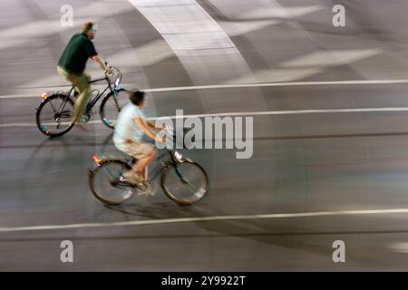 Dynamische Schwenkaufnahme für zwei Radfahrer, die nachts in der geschäftigen Stadt fahren. Bewegung und Geschwindigkeit betonen das urbane Nachtleben. Stockfoto