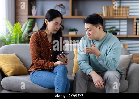 Asiatisches Paar, das auf der Couch sitzt und sich ernsthaft unterhalten hat. Frau, die das Telefon hält, Mann erklärt es. Szene veranschaulicht Kommunikation und Technologie in der modernen Interaktion mit der Familie. Stockfoto