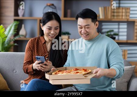 Asiatisches Paar genießt Pizza-Liefererfahrung und teilt Glück zu Hause. Die Frau hält das Telefon, während der Mann die Pizzaschachtel öffnet und in ungezwungener Atmosphäre Freude und Zweisamkeit zum Ausdruck bringt. Stockfoto