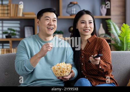 Asiatisches Paar lächelt beim Fernsehen zu Hause und teilt Popcorn auf dem Sofa. Drückt Freude, Entspannung und gemeinsame Freizeit aus. Fernbedienung in der Hand für komfortables Entertainment-Erlebnis. Stockfoto