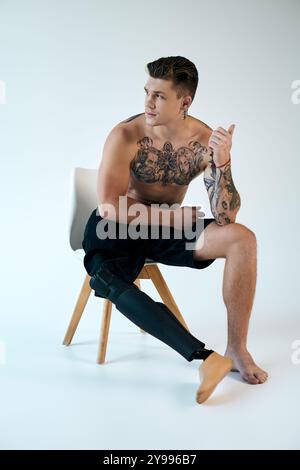 Ein junger Mann mit Tattoos sitzt auf einem Stuhl und zeigt selbstbewusst seine Beinprothese in einem eleganten Studio. Stockfoto