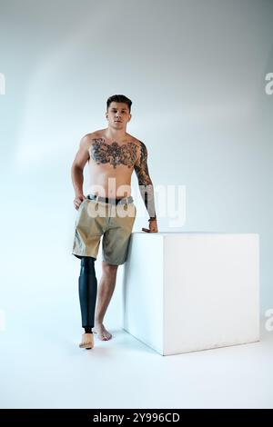 Ein selbstbewusster junger Mann mit Tattoos steht stolz in einem Studio und zeigt seine Beinprothese und seine Kraft. Stockfoto